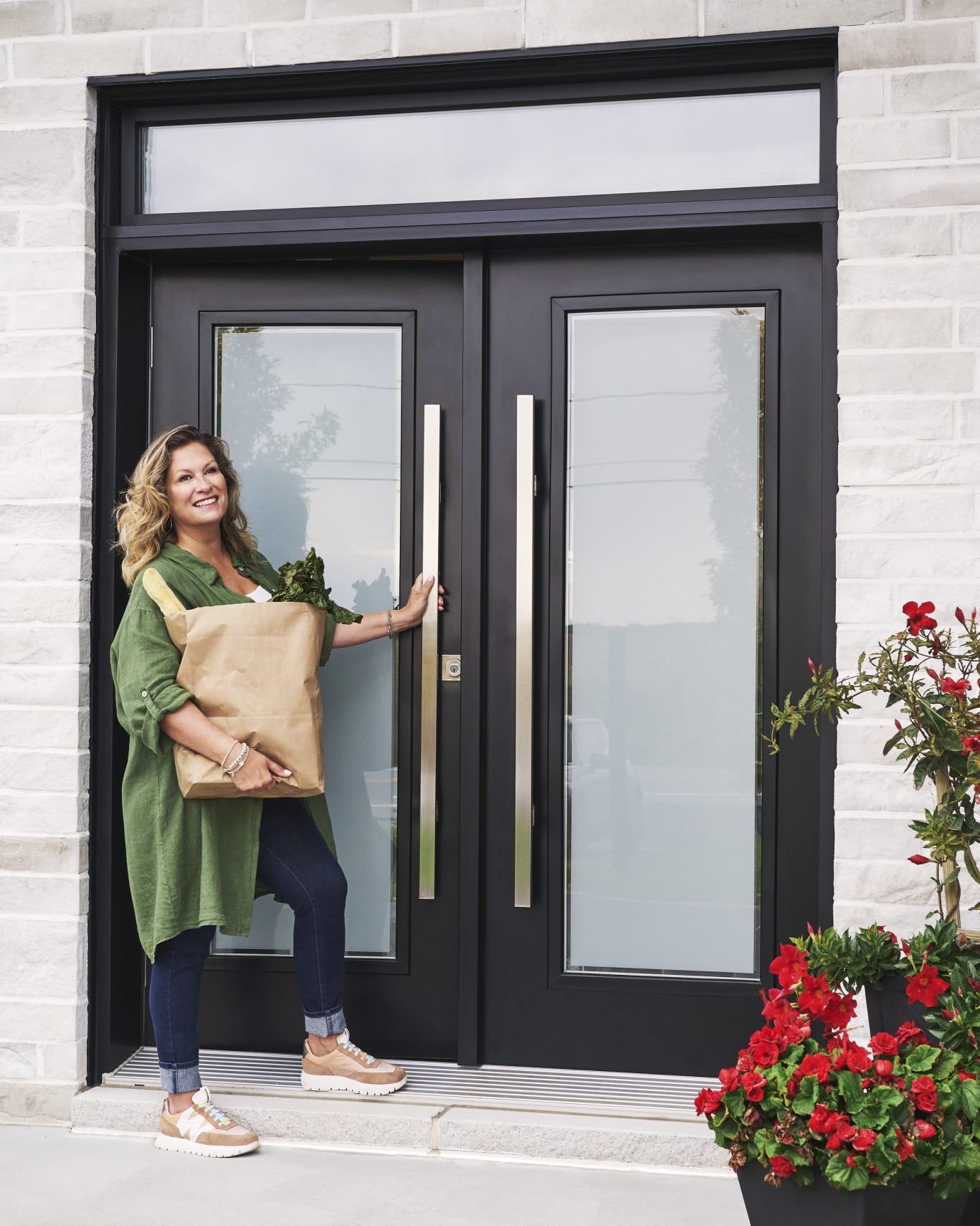 Saskia Thuot, un sac d'épicerie dans les bras, ouvre une grande porte d'entrée Isothermic noire à deux vantaux vitrés, dans une façade de pierre, près de fleurs rouges