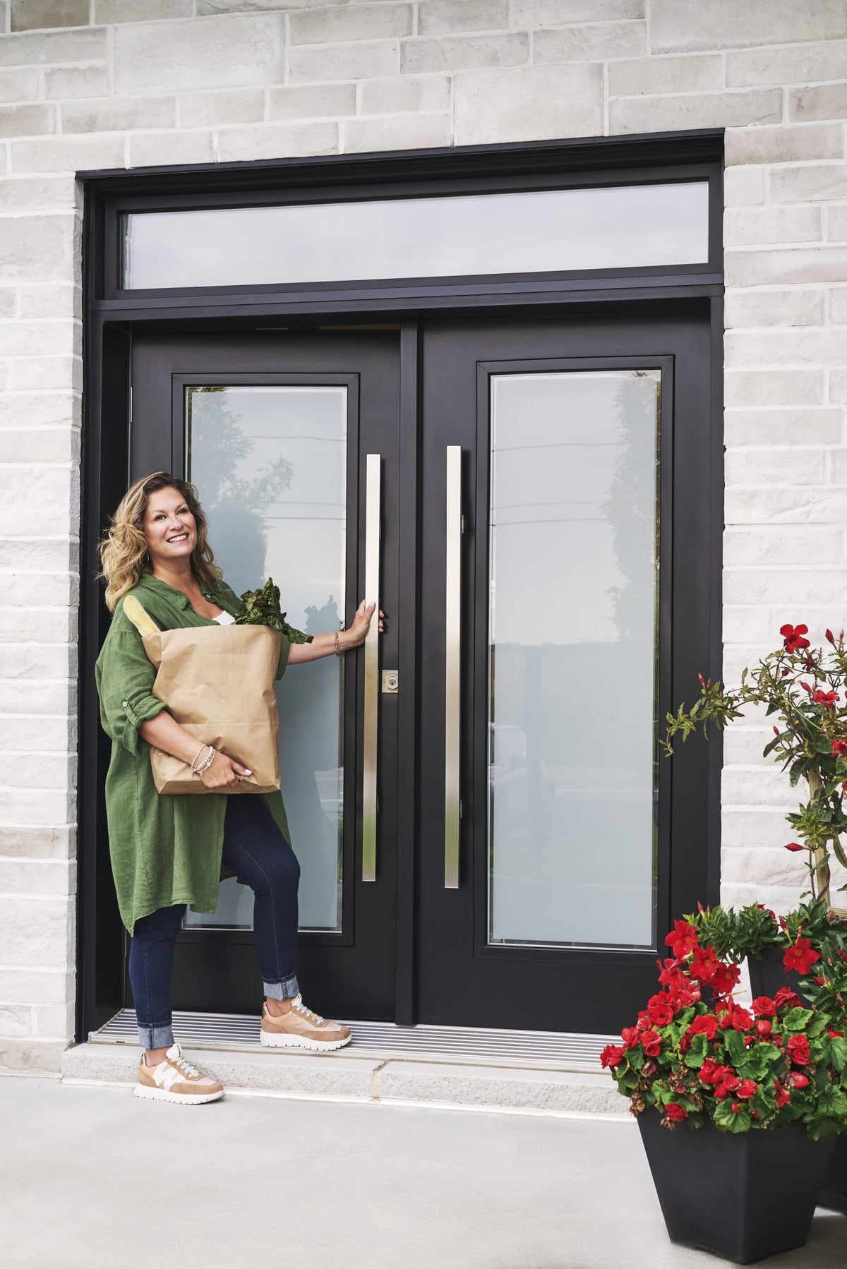 Saskia Thuot, un sac d'épicerie dans les bras, ouvre une grande porte d'entrée Isothermic noire à deux vantaux vitrés, dans une façade de pierre, près de fleurs rouges