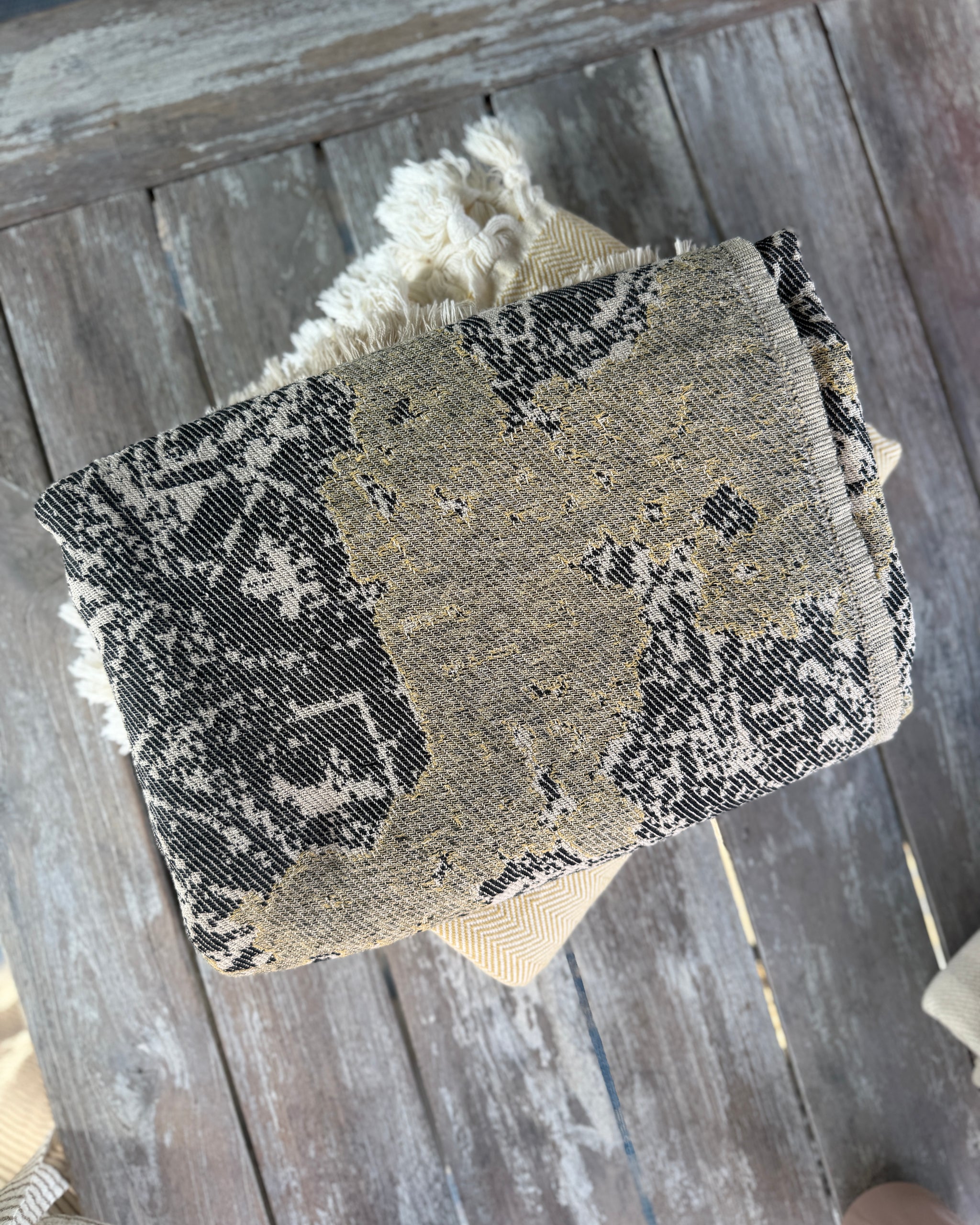 Jeté GEORGIE roulé sur une table en bois : jacquard noir et or, franges crème