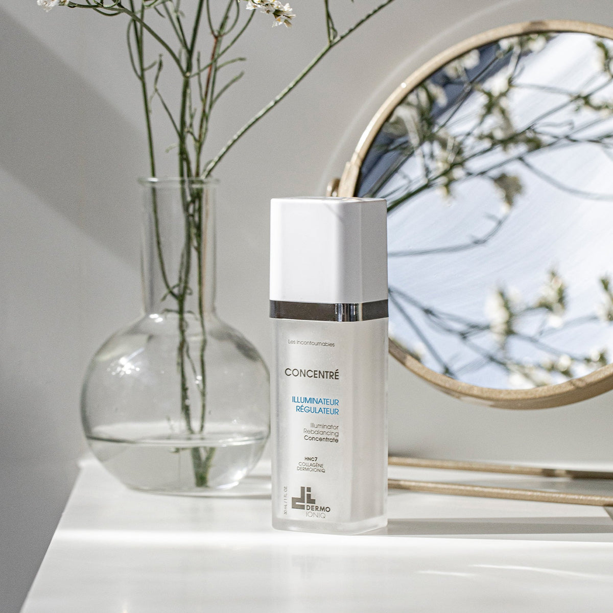 Flacon givré Dermo Ioniq Concentré Illuminateur Régulateur sur tablette blanche, près d’un vase en verre et d’un miroir rond